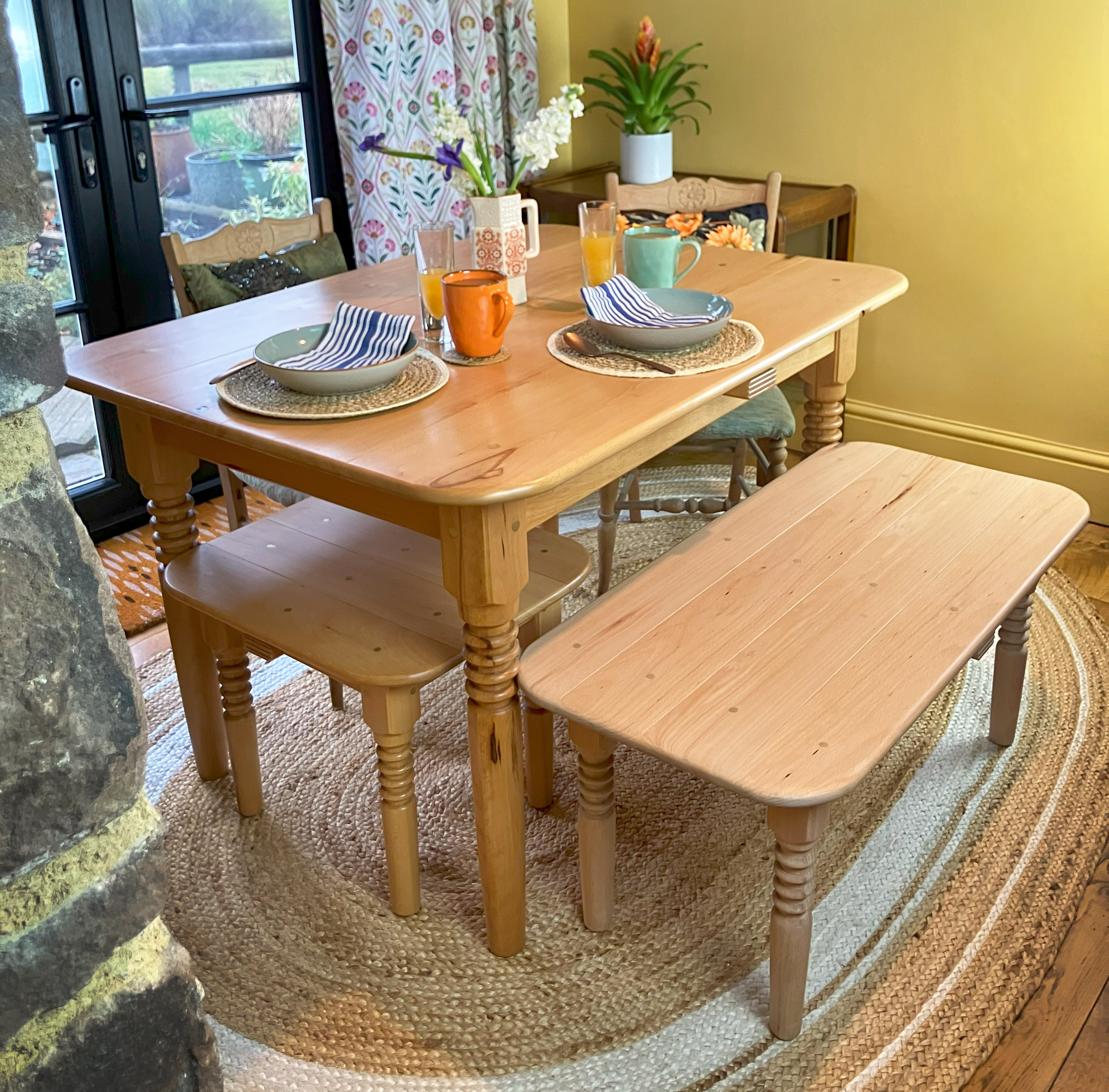 Solid wood handmade contemporary farmhouse table with unique turned legs and brass dowels, set for breakfast in a rustic setting with matching bench and seat beside it.