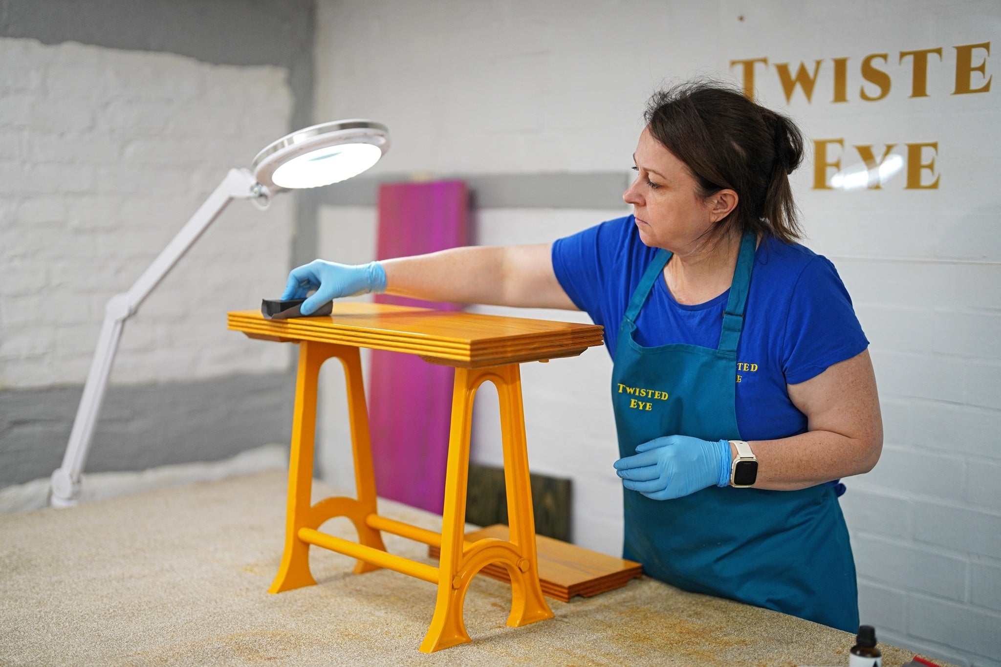 Person in a blue apron working on a yellow stool with a magnifying lamp in a workshop setting.